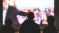 Eine Leinwand mit projeziertem Film davor stehen drei Personen, die auf die Leinwand schauen