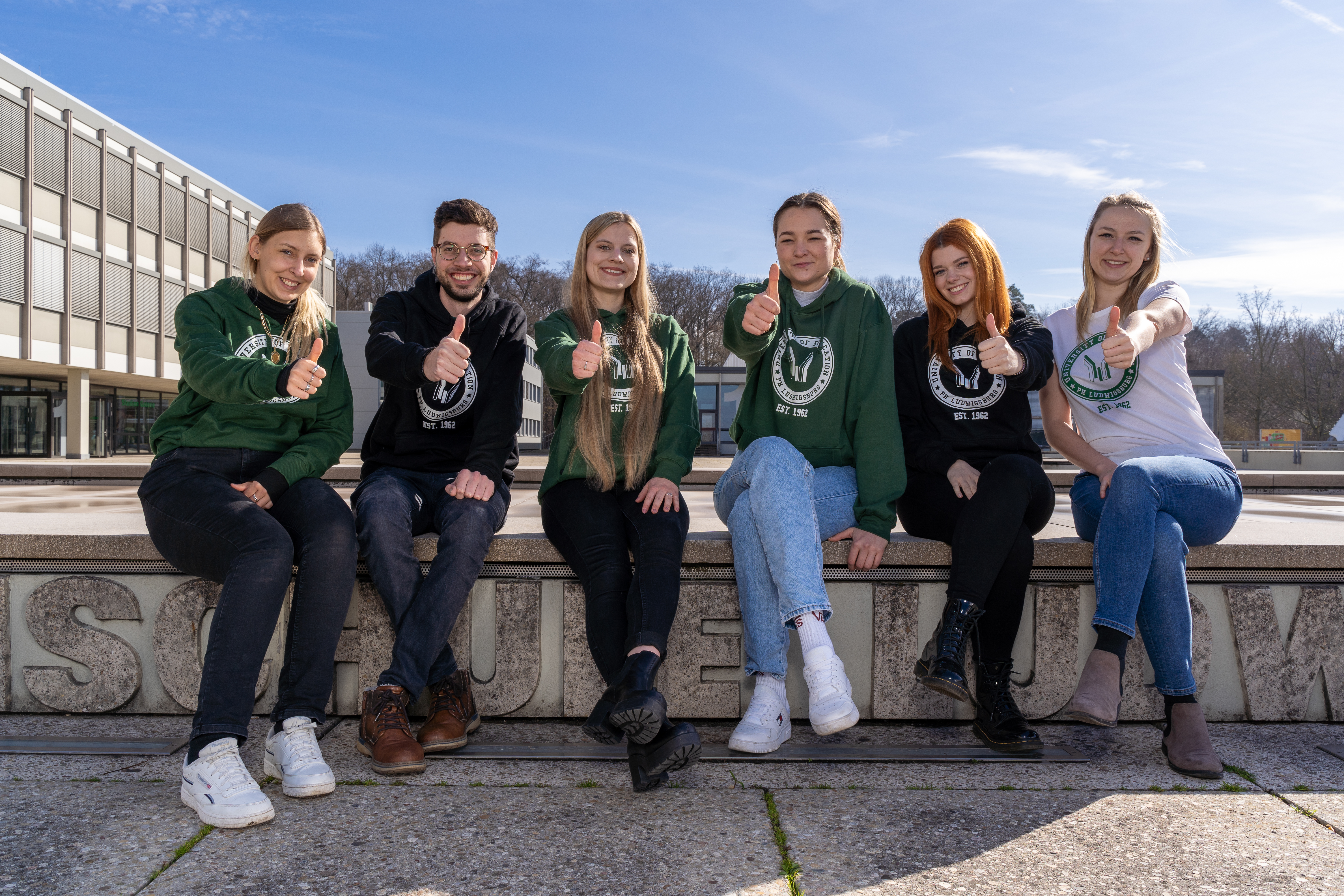Lächelnde Studierende sitzen auf der Umrandung des Brunnens der PH Ludwigsburg und zeigen mit den Daumen nach oben