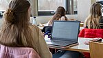 Blick von hinten auf Studentinnen, die in einem Seminarraum einem Dozierenden zuhören. Auf dem Tisch vor der einer Studentin steht ein aufgeklapptes Laptop.
