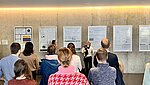 Das Bild wurde bei einer Posterpr&auml;sentation aufgenommen. Man sieht eine Person, die ein Poster vorstellt, und mehrere Personen mit dem R&uuml;cken zur Kamera, die der pr&auml;sentierenden Person zuh&ouml;ren. 