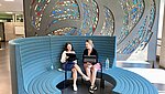 Zwei Studentinnen sitzend auf einem blauen Rundsofa, das vor einer Wand mit Mosaikglas steht. 
