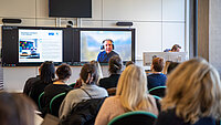 Das Foto wurde beim Forschungstag 2024 aufgenommen. Zu sehen ist der Vortragende Prof. Dr. Pinkwart auf dem Bildschirm, das Publikum ist von hinten zusehen und blickt auf den Monitor.