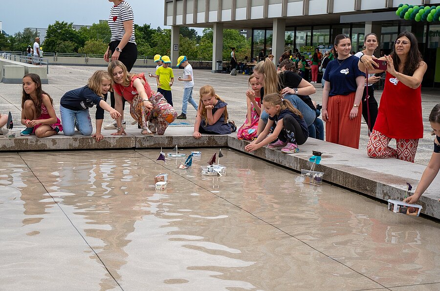 Kinder sitzen um den Brunnen der PH Ludwigsburg und lassen selbstgebasteltete Boote ins Wasser