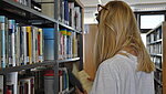 Frau steht vor einem B&uuml;cherregal, mit dem R&uuml;cken zum Betrachter