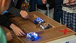 Mehrere Kinder stehen um ein Tischlabyrinth, in dem drei kleine, blau leuchtende Lernroboter (Blue-Bots) navigiert werden.