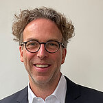 Herr Sven Wippermann hat braune Locken. Er tr&auml;gt ein schwarzes Jacket und ein wei&szlig;es Hemd. 