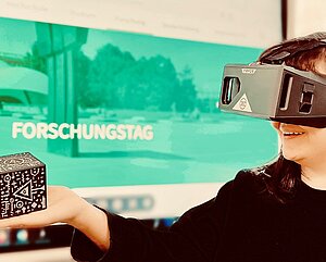 Eine Forscherin trägt eine VR-Brille und schaut auf ein Spielobjekt. Hinter ist ein Bildschirm mit der Folie Forschungstag