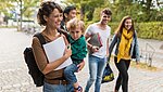 Studierende lachend und gehend auf dem Campus. Im Vordergrund eine junge Studentin mit Kind auf dem Arm