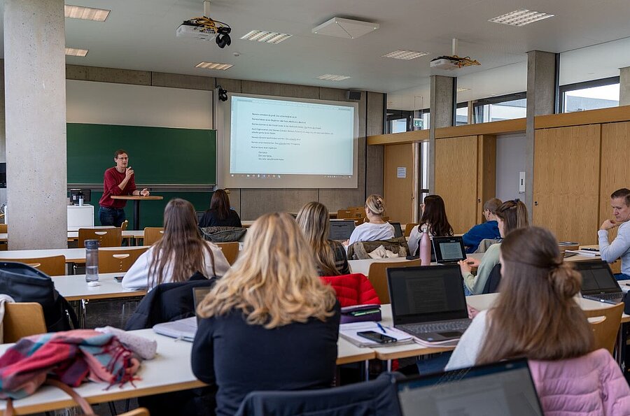 Blick auf hinten auf Studentinnen und Studenten, die einem vortragenden Dozierendem zuhören. Im Hintergrund wird auf eine Projektionsfläche eine Folie gezeigt.