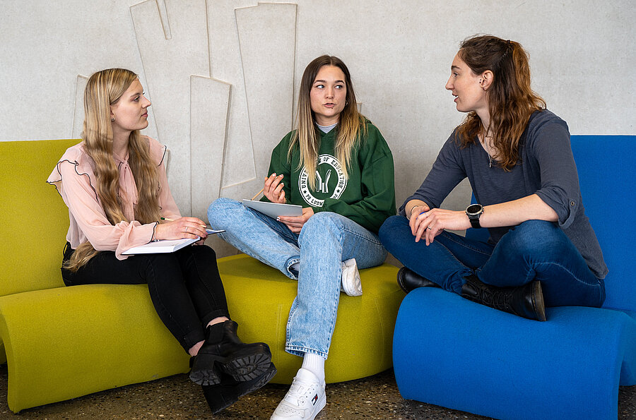 drei Studentinnen sitzen auf den bunten Sitzhockern des Lichtgangs der Pädagogischen Hochschule Ludwigsburg.