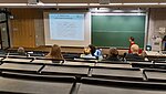 Vorlesungssituation in einem Hörsaal. Der Blick richtet sich von hinten nach vorne die Hinterköpfe der Studierenden, den Dozierenden und die Projektiosfläche sowie die Schreibtafeln. 